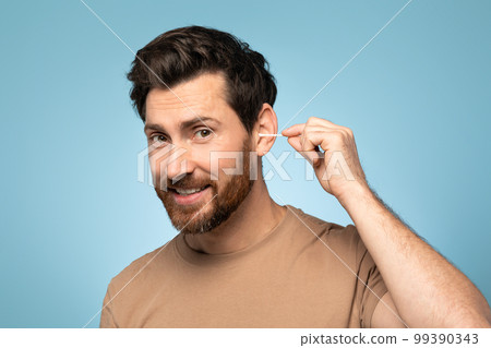 Happy handsome man using cotton swab stick for ear clean, posing over blue background and smiling at camera 99390343
