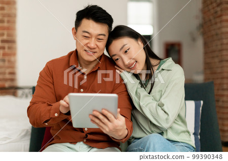 Happy korean spouses using digital pad, surfing internet or shopping online, sitting on sofa at home, free space Happy korean spouses using digital pad, surfing internet or shopping online, sitting on sofa at home, free space 99390348