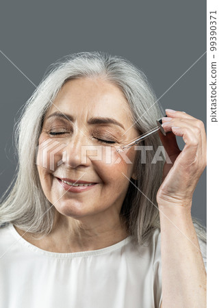 Smiling old european female with gray hair applies serum, oil on her face, isolated on gray background 99390371