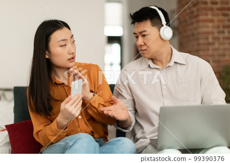Marital problem. Asian middle aged husband looking at wife's smartphone while using laptop and wearing headphones 99390376