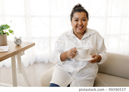 Positive young chubby woman sitting at workdesk, drinking coffee Positive young chubby woman sitting at workdesk, drinking coffee 99390537