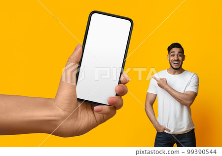 Cheerful arab man pointing at big blank smartphone in giant female hand Cheerful arab man pointing at big blank smartphone in giant female hand 99390544