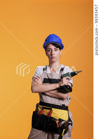 Professional handywoman holding electric power drill, wearing hardhat and overalls to renovate walls. Serious construction worker using nail drilling gun, working tool on studio background. 99393457