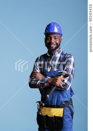 Happy African american builder holding hammer posing in studio against blue background. Workman in hard hat and construction overalls, safety uniform in construction industry. Happy African american builder holding hammer posing in studio against blue background. Workman in hard hat and construction overalls, safety uniform in construction industry. 99393458