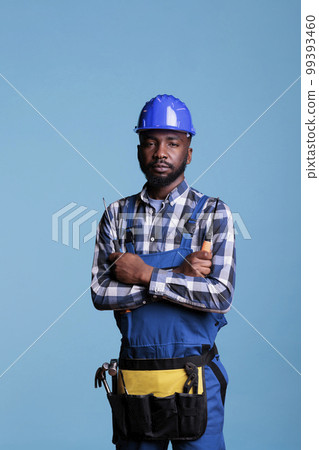 Portrait of construction professional in front of camera holding screwdriver in studio shot. African american builder wearing tool belt and work uniform isolated on blue background. 99393460