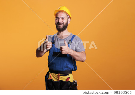 Successful repairman giving thumbs up on camera, showing like and approval gesture in studio. Male construction worker expressing agreement and good symbol, confident happy expert. 99393505