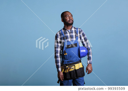 African american construction worker thoughtful looking into distance while holding hard hat under arm. Contractor wearing work clothes and tools against blue background, studio shot. 99393509