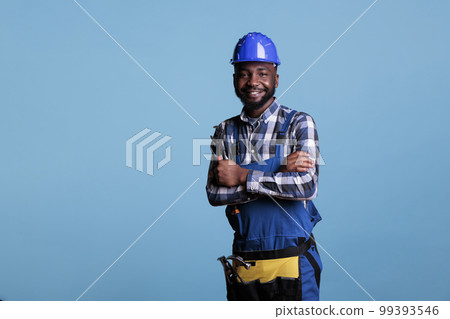 African american construction worker with happy face wearing a helmet smiling while looking at camera. Confident builder posing with arms crossed holding wrench against blue background. 99393546