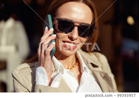 Smiling woman in sunglasses is talking phone on city street background and looking at camera Smiling woman in sunglasses is talking phone on city street background and looking at camera 99393957