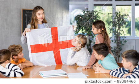 Schoolchildren listen carefully to story about the country England. Teacher holds flag of England in her hands Schoolchildren listen carefully to story about the country England. Teacher holds flag of England in her hands 99394466
