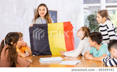 Positive young woman teacher showing flag of Belgium to schoolchildren preteens during history lesson in audience 99394543