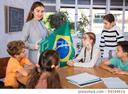 Happy young woman teacher conducting lesson for preteens and showing state flag of Brazil in audience of middle school Happy young woman teacher conducting lesson for preteens and showing state flag of Brazil in audience of middle school 99394619