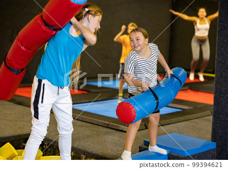 Smiling positive girls in sportswear having funny wrestling by inflatable logs in indoor game trampoline club Smiling positive girls in sportswear having funny wrestling by inflatable logs in indoor game trampoline club 99394621