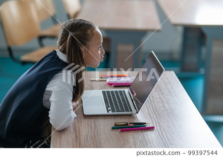 Caucasian girl communicates via video communication on a laptop while sitting at a school desk.  99397544