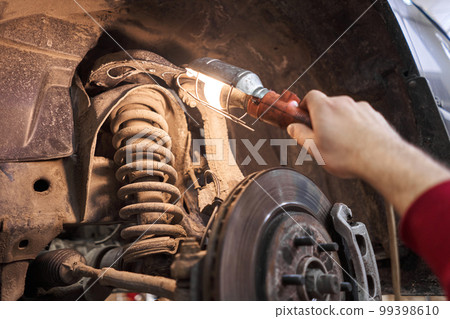 A car mechanic inspects springs, shock absorbers and suspension with a flashlight. Car on a repair stand. Technical service station for car. A car mechanic inspects springs, shock absorbers and suspension with a flashlight. Car on a repair stand. Technical service station for car. 99398610