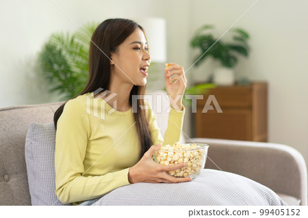 Young asian woman putting a bowl of popcorn on pillow and eating popcorn while sitting on the big comfortable 99405152