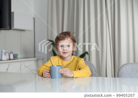Happy cute child boy in yellow sweater sitting by the table and holding mug  99405207