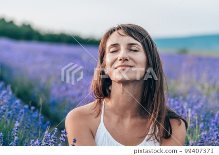 Close up portrait of happy young brunette woman in white dress on blooming fragrant lavender fields with endless rows. Warm sunset light. Bushes of lavender purple aromatic flowers on lavender fields. Close up portrait of happy young brunette woman in white dress on blooming fragrant lavender fields with endless rows. Warm sunset light. Bushes of lavender purple aromatic flowers on lavender fields. 99407371