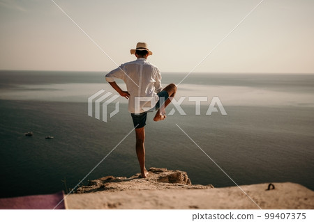 A digital nomad, a man in a hat, a businessman does yoga on the rocks by the sea at sunset time, does a business operation online from a distance. Remote work on vacation. A digital nomad, a man in a hat, a businessman does yoga on the rocks by the sea at sunset time, does a business operation online from a distance. Remote work on vacation. 99407375