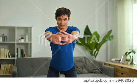 Handsome sporty man exercising and doing squats in living room. Sport and healthy lifestyle concept Handsome sporty man exercising and doing squats in living room. Sport and healthy lifestyle concept 99408020