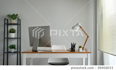 Wooden table with computer monitor and stationery and lamp in cozy home office interior decorated with green potted plants 99408033
