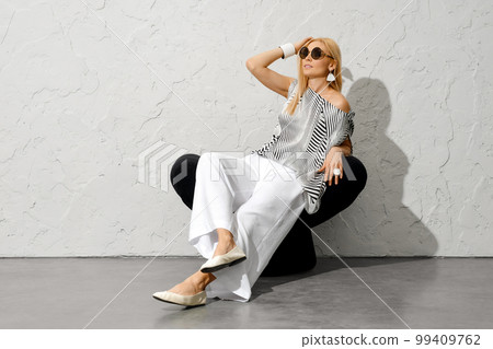 Charming woman in sunglasses, striped blouse and wide trousers sitting on armchair 99409762