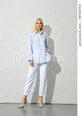 Woman in white pants, stilettos and blue cotton blouse posing near white wall 99409763