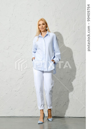 Trendy woman in white pants and blue cotton blouse moves away from white wall Trendy woman in white pants and blue cotton blouse moves away from white wall 99409764
