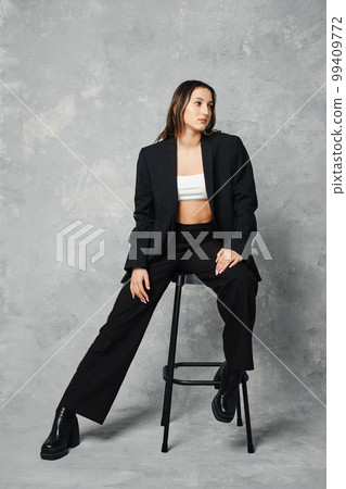 Young woman in black trendy pantsuit and ankle boots with rough sole sitting on tall stool Young woman in black trendy pantsuit and ankle boots with rough sole sitting on tall stool 99409772