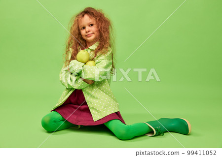 Vitamins and taste. Little cute girl, child with curly hair posing with apples over green studio background. Concept of childhood, emotions, fun Vitamins and taste. Little cute girl, child with curly hair posing with apples over green studio background. Concept of childhood, emotions, fun 99411052