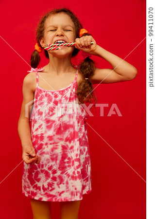 Emotionally eating candy. Little cute girl, child with curly hair posing over bright red studio background. Concept of childhood, emotions, fun Emotionally eating candy. Little cute girl, child with curly hair posing over bright red studio background. Concept of childhood, emotions, fun 99411070
