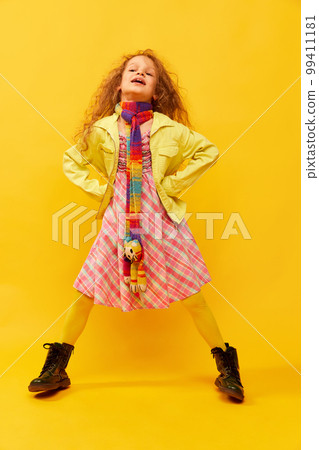 Extraordinary look. Little cute girl, child with curly hair posing in bright clothes over yellow studio background. Concept of childhood, emotions, fun 99411181