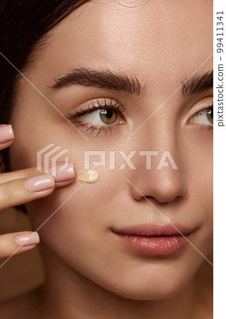 Close-up portrait of beautiful young brunette girl with perfect skin applying cream foundation on face over brown studio background. Concept of natural beauty, skincare, cosmetology 99411341