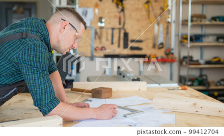 A portrait of a carpenter in goggles and work overalls draws a blueprint for a workshop. 99412704