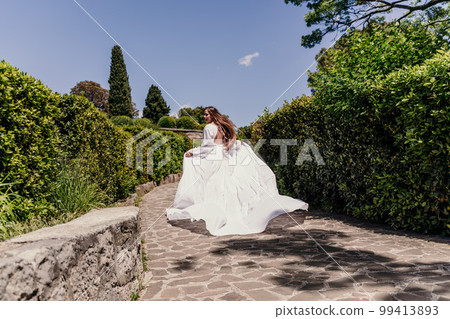 Brunette runs white dress park. A beautiful woman with long brown hair and a long white dress runs along the path along the beautiful bushes in the park, rear view Brunette runs white dress park. A beautiful woman with long brown hair and a long white dress runs along the path along the beautiful bushes in the park, rear view 99413893