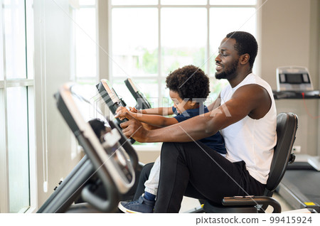African American man with child exercising on the exercise bike in indoor fitness club. Healthy and Fitness concept. African American man with child exercising on the exercise bike in indoor fitness club. Healthy and Fitness concept. 99415924