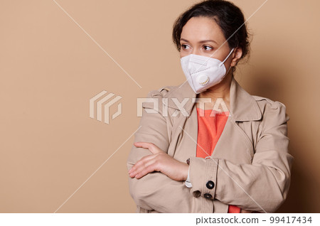 Portrait of confident business woman in safety medical mask, looking aside at copy advertising space, posing with her arms folded on isolated beige background. Social distancing and quarantine concept 99417434