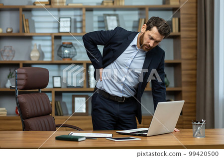 Mature man working in modern office with laptop, senior boss having severe back pain, businessman at workplace overworked working late massaging his back with hand, severe pain from sitting. Mature man working in modern office with laptop, senior boss having severe back pain, businessman at workplace overworked working late massaging his back with hand, severe pain from sitting. 99420500