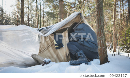 A homeless man climbs in and out of a tent in the woods in winter. A homeless man climbs in and out of a tent in the woods in winter. 99422117