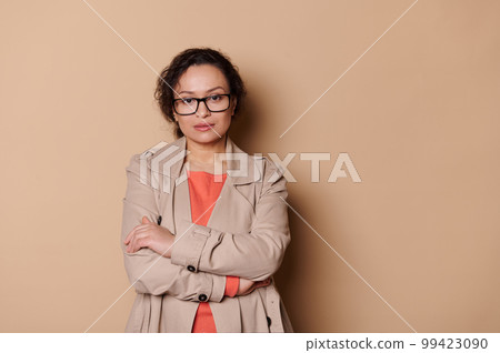 Confident portrait of a multi-ethnic beautiful middle-aged woman, wearing stylish eyeglasses, confidently looking at camera, isolated on beige color background. Copy advertising space. 99423090