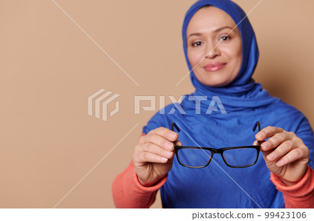 Selective focus on black spectacles - eyeglasses in the hands of a blurred stylish Arab Muslim in blue hijab, smiling looking at camera, isolated on beige background. Health Fashion and ophthalmology 99423106