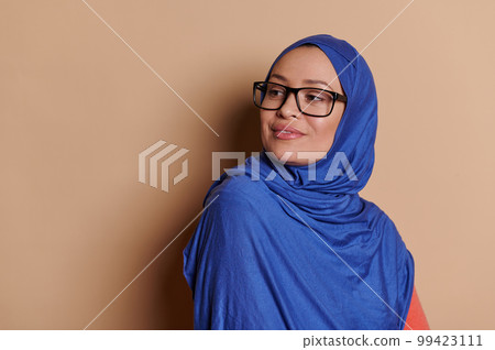 Attractive confident Middle-Eastern Muslim woman in blue hijab, squinting looking to the side through her stylish eyeglasses, isolated on cream background. Vision problems concept. Copy space for ads Attractive confident Middle-Eastern Muslim woman in blue hijab, squinting looking to the side through her stylish eyeglasses, isolated on cream background. Vision problems concept. Copy space for ads 99423111
