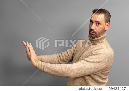 Side view of serious man making stop gesture showing palm, conflict ban warning about danger, stop bullying, wears beige turtleneck sweater. Indoor studio shot isolated on gray background 99423417