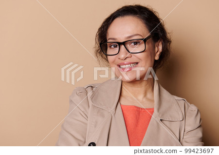 Closeup portrait of a middle-aged ethnic pretty woman in beige coat, wearing stylish eyeglasses, smiling with a beautiful a toothy smile looking at camera, isolated on cream background. Ophthalmology 99423697