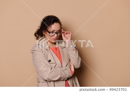 Isolated portrait on beige background of a multi-ethnic middle-aged curious brunette lady, holding stylish spectacles, looking at empty space for advertising text. Health. Fashion. Eyesight problems 99423701