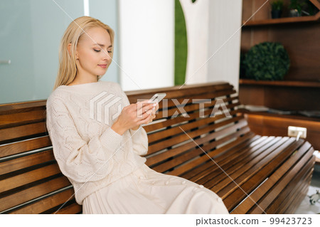 Medium shot of young blonde woman sitting on wooden bench using smartphone looking at cellphone screen enjoying using mobile apps. Happy female checking social media holding smartphone. 99423723