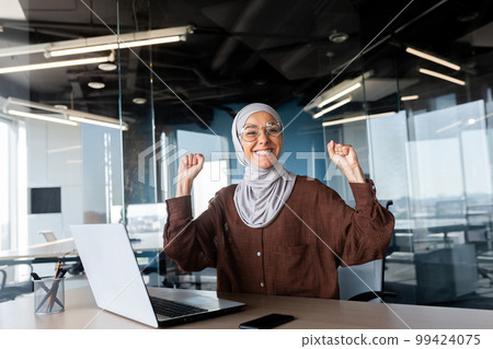Portrait of happy smiling and successful muslim at work in office, businesswoman with laptop holding hands up gesture of success and triumph, woman in hijab received online victory notification. 99424075