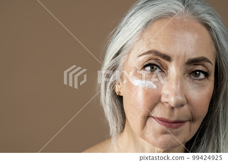 Calm old european lady with gray hair applies moisturizer on face, isolated on brown background, close up, studio 99424925