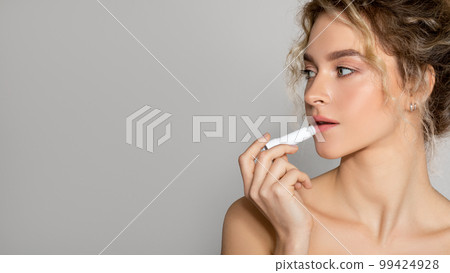 Young beautiful woman with perfect skin applying hygenic lipbalm on lips, looking aside at free space, grey background 99424928