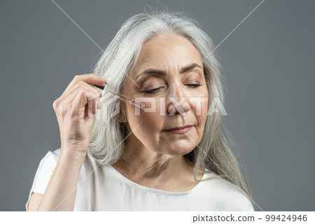 Calm old european lady with gray hair applies serum, oil on her face, enjoy procedures 99424946